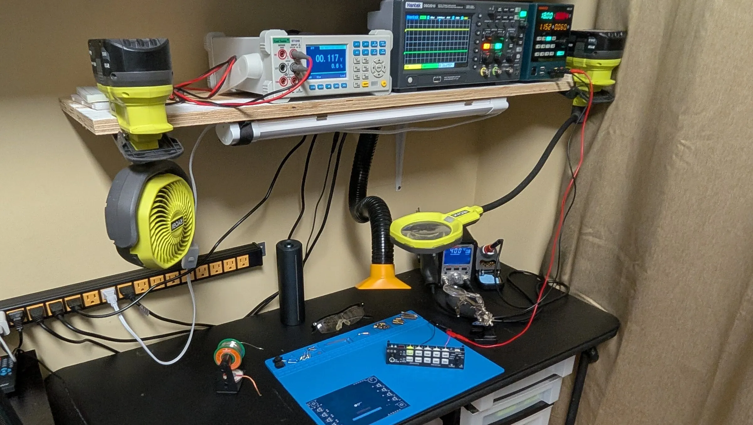 A soldering and testing bench with an oscilloscope, multimeter and power supply on a shelf above an audio panel prototype